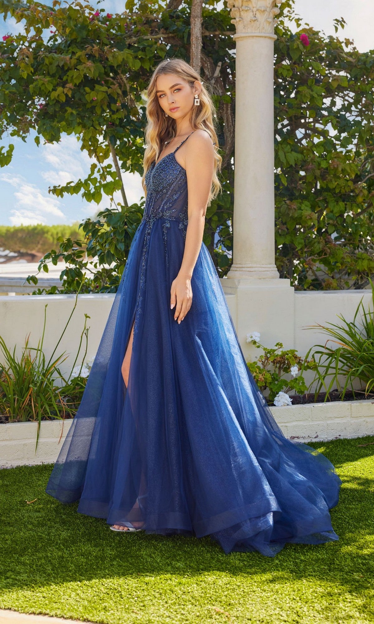 A woman stands outdoors in the Juliet JT2632L long prom dress, a sleeveless blue A-line gown with beaded embroidery, sheer overlay, and slit, framed by greenery and white columns.